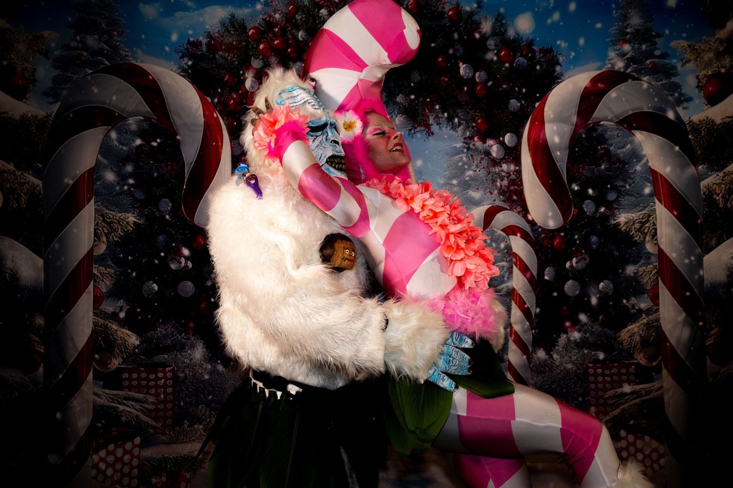 Two performers in festive candy cane Christmas costumes pose against a holiday backdrop with ornaments and snow.