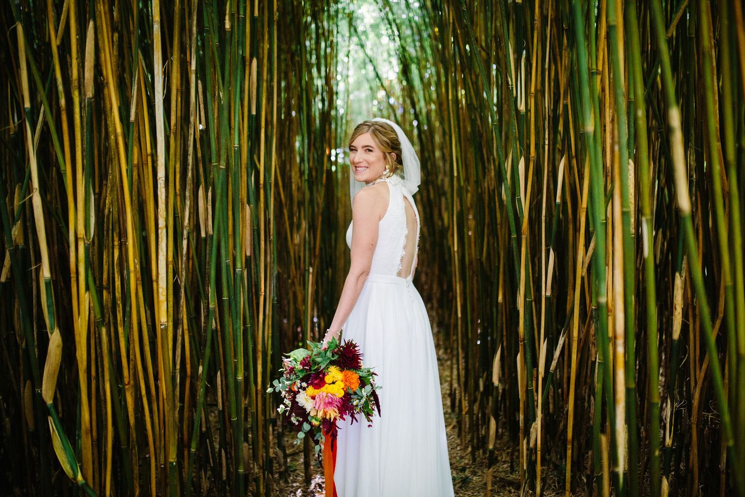 Bride in white gown holding colorful bouquet stands in lush bamboo forest, smiling over her shoulder.