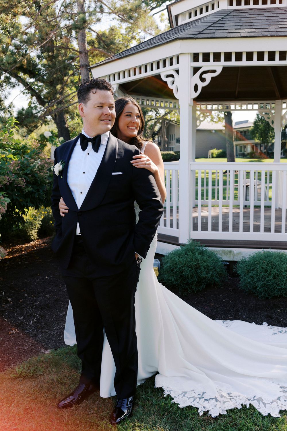 Bride and groom pose together in front of a white gazebo in a lush Midwest garden setting.