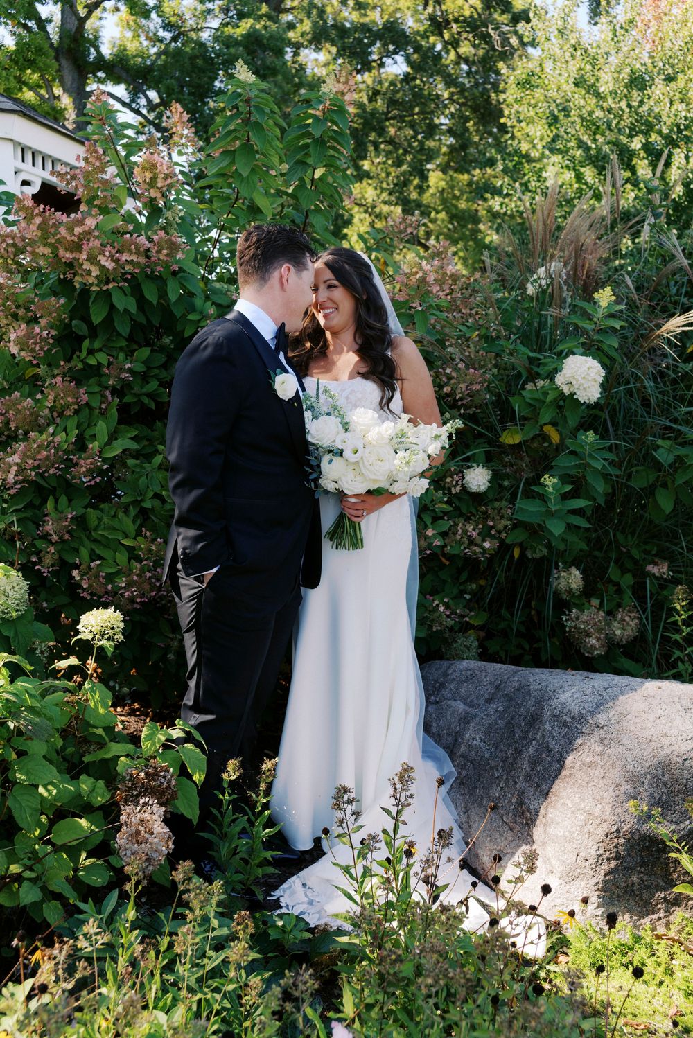 Bride and groom share a romantic moment surrounded by lush garden greenery, bride holding white floral bouquet.