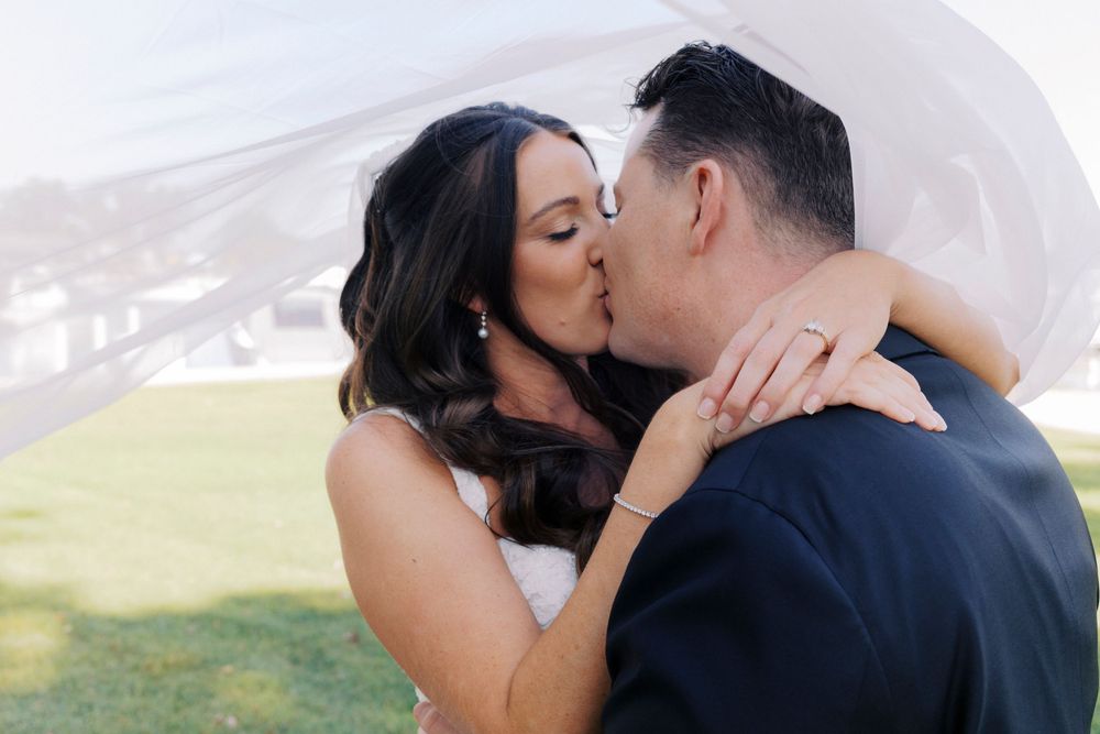 Bride and groom sharing a romantic kiss under a flowing veil on their wedding day outdoors.
