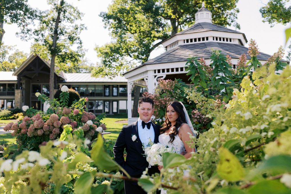 Bride and groom pose among lush garden greenery with elegant Abbey Resort in background on sunny wedding day.