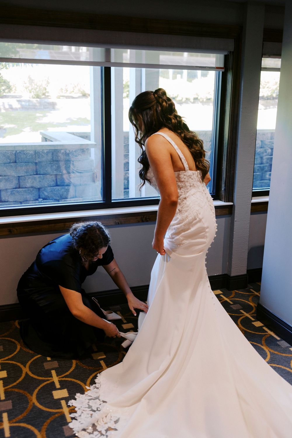 Bride in white mermaid wedding gown being helped by assistant near large windows before ceremony.