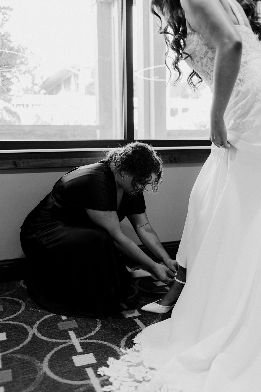 Bridesmaid kneeling to help bride put on white heels before wedding ceremony, black and white photo.