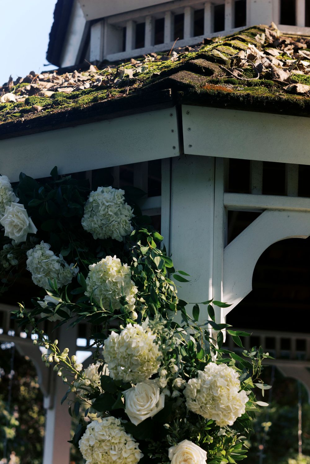 White hydrangeas and roses adorn a gazebo corner with lush greenery against a moss-covered tile roof.