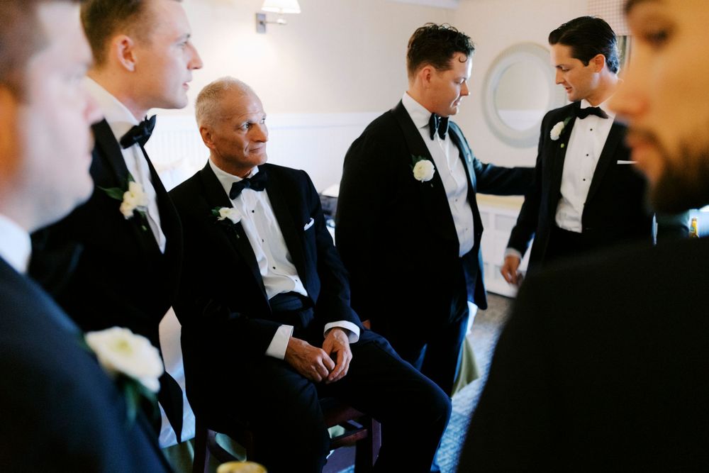 Groomsmen in black tuxedos with bow ties gather before a wedding ceremony in an elegant white venue.