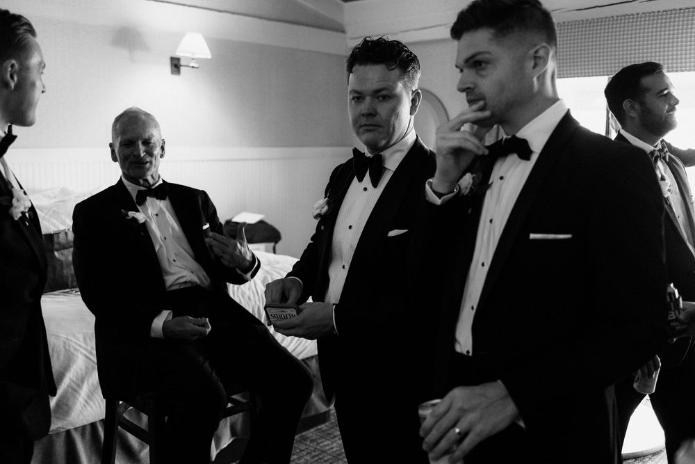 Black and white photo of groomsmen in tuxedos preparing together before a wedding ceremony.