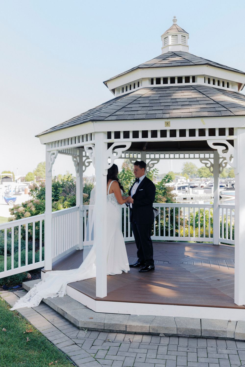 Bride and groom share a romantic moment in a white gazebo overlooking a waterfront marina on their wedding day.