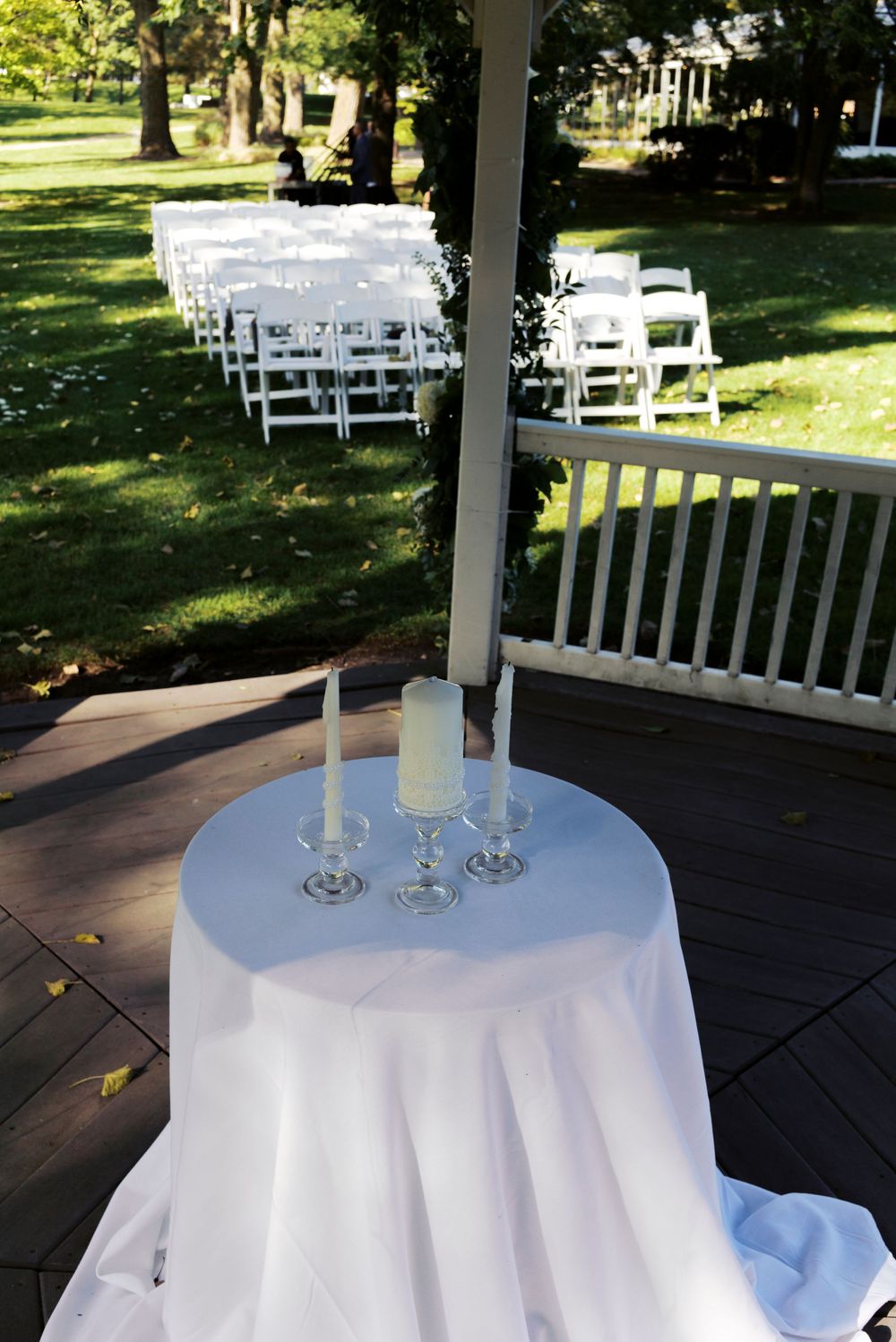 Outdoor wedding ceremony setup with white chairs on green lawn, unity candle table with white tablecloth on wooden deck.
