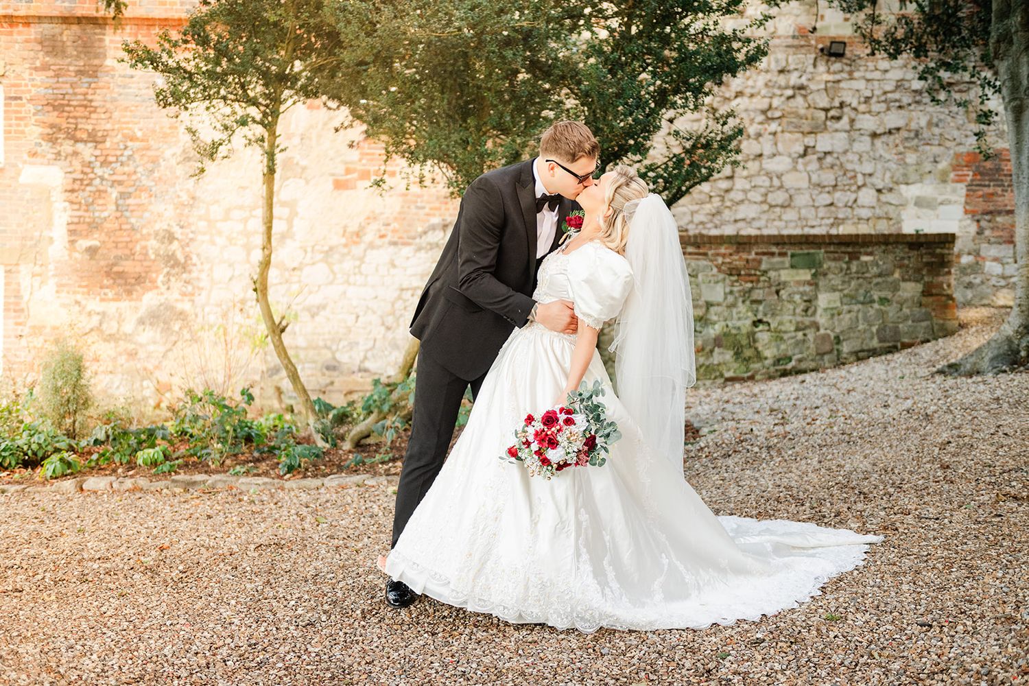 Bride and groom sharing a romantic kiss outdoors, bride holding red bouquet in elegant white gown with long veil.
