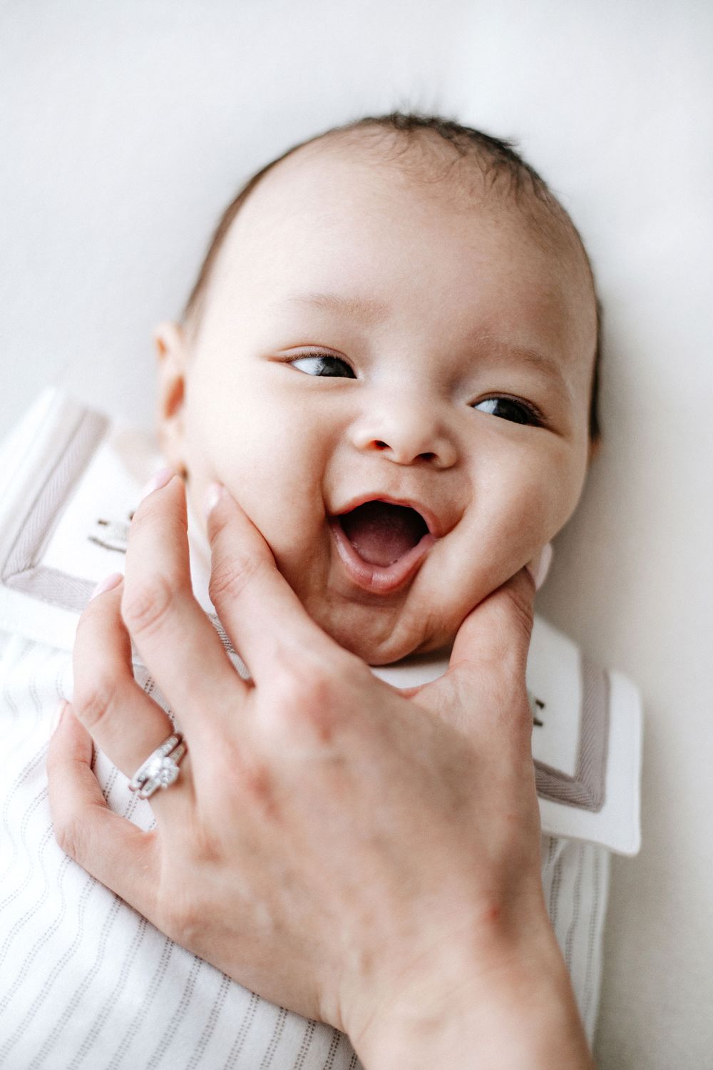 An adorable baby with a wide joyful smile while being held by mothers hand wearing a ring.