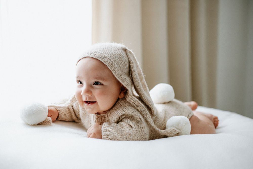 Baby in cosy knit hoodie with pom poms lies on white bedding while smiling.