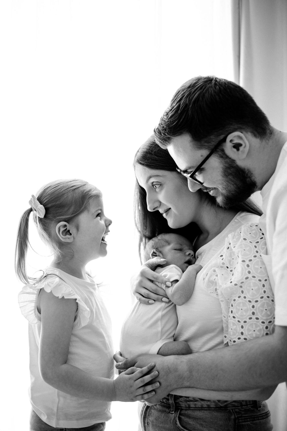 Black and white portrait of a family looking lovingly at their newborn baby while young daughter watches.