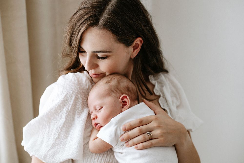 A tender moment of bonding between parent and sleeping infant in white clothing by a window.