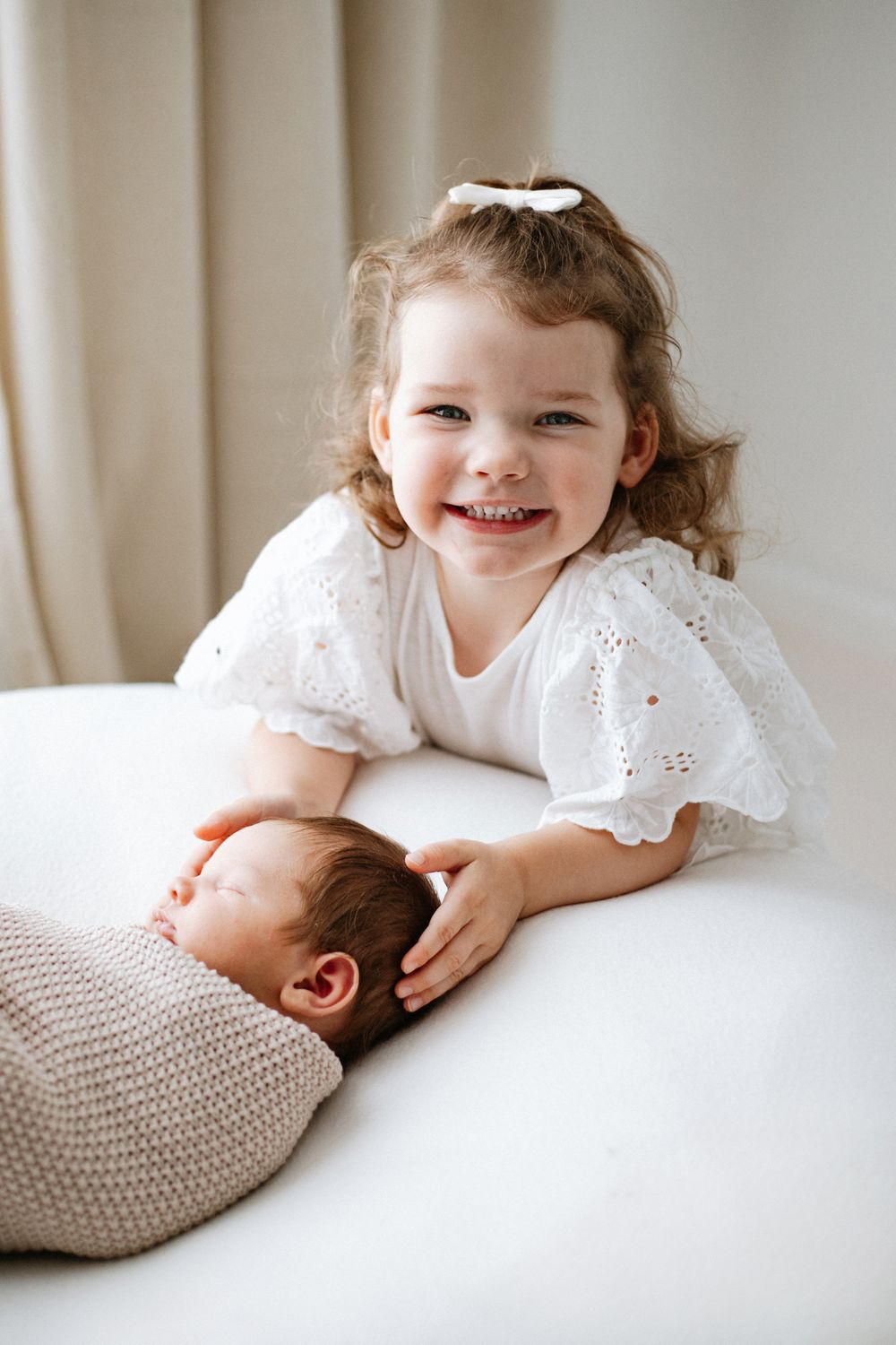 Child in white lace dress smiling while petting a sleeping baby's head on white bedding.
