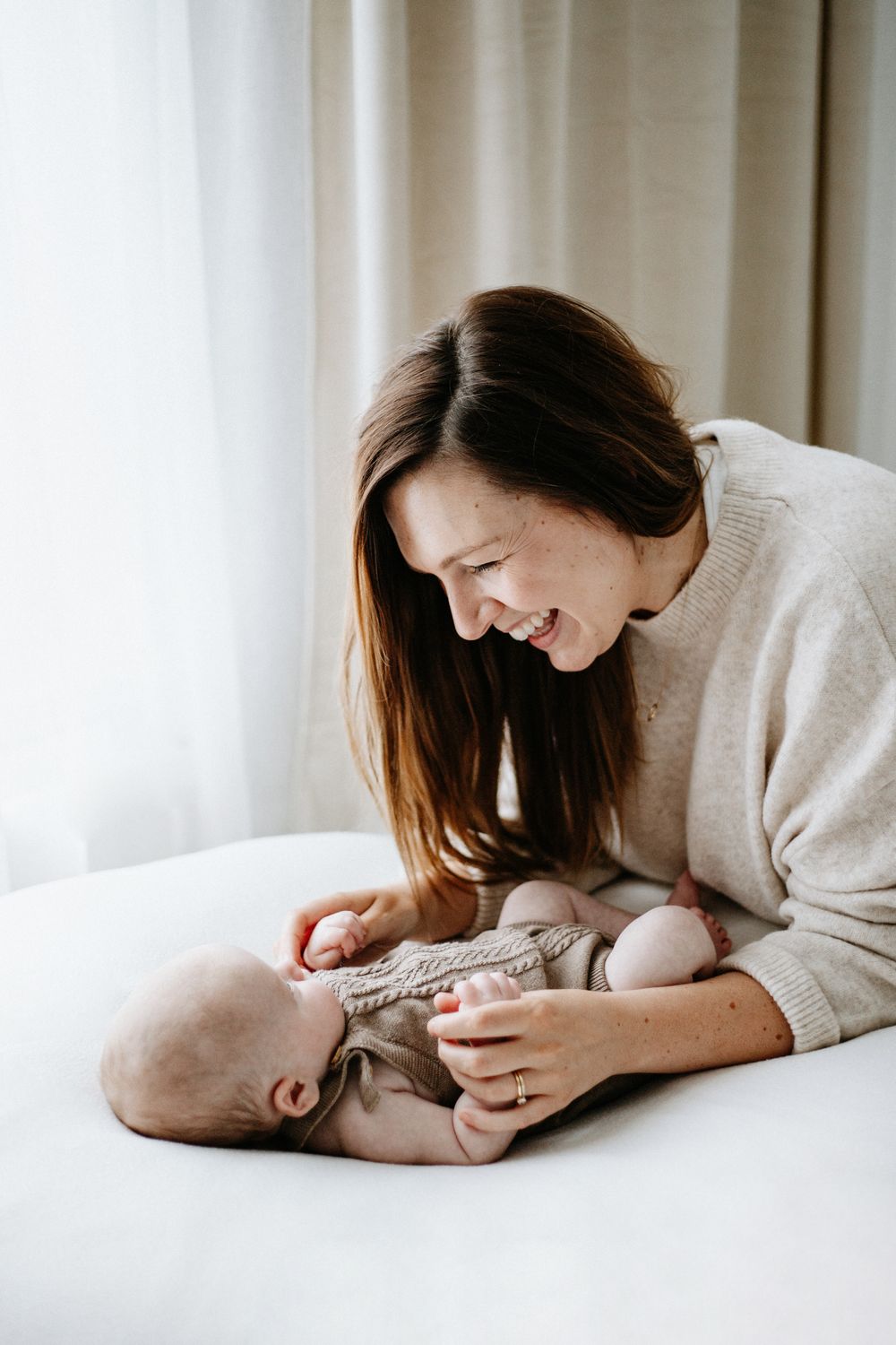 Parent in cozy white sweater sharing a tender moment with baby lying on white bedding.