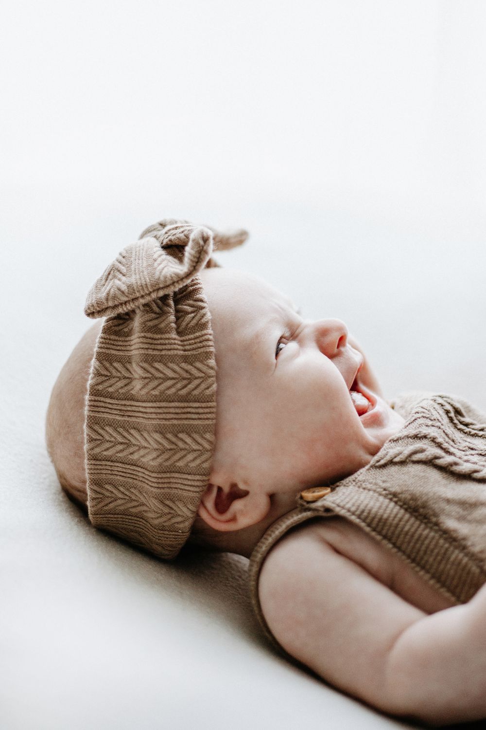 A baby wearing a tan patterned headband laughs while lying down on a newborn posing beanbag.