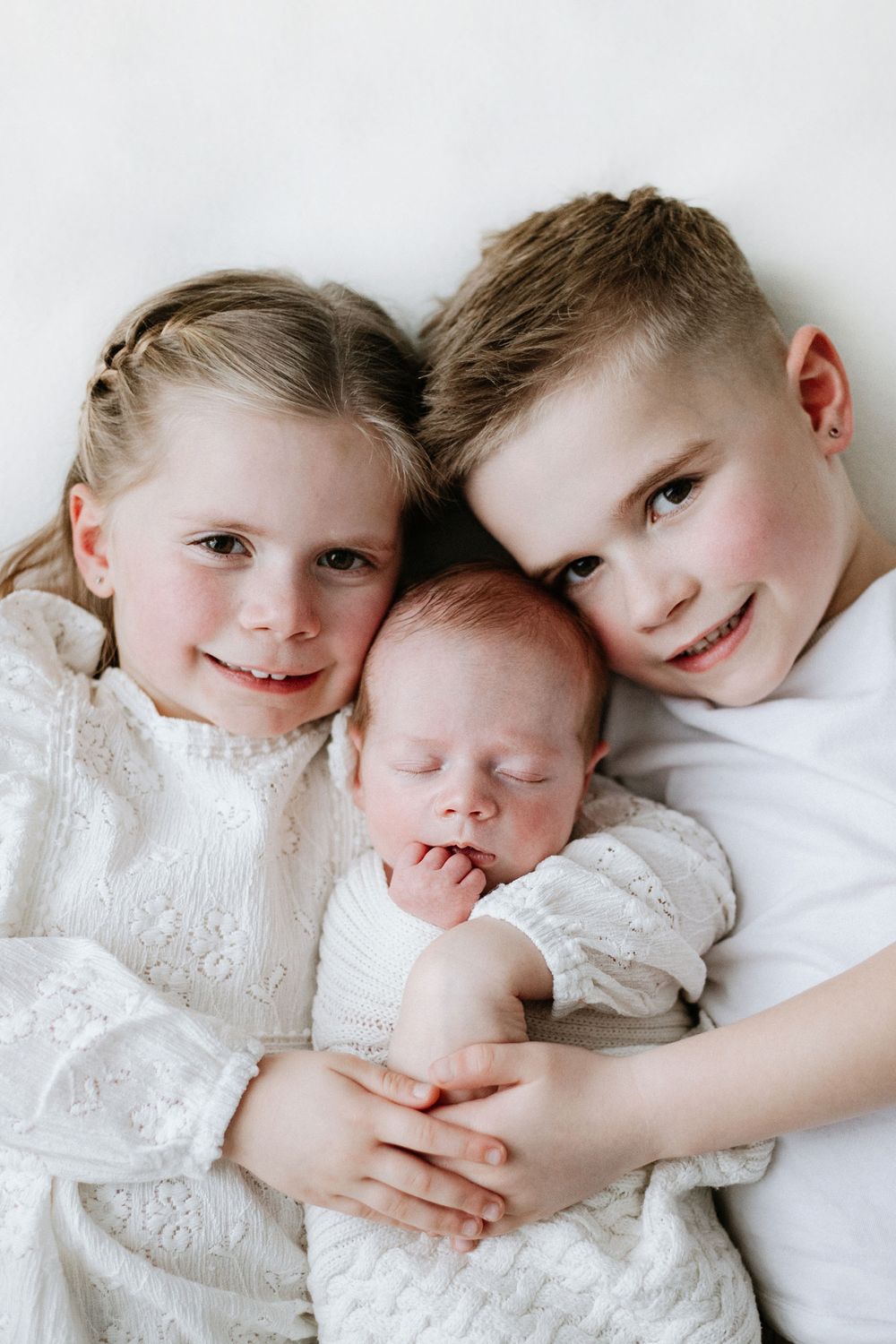 Three siblings in white clothing, including a baby and two older children, snuggle close together in a tender portrait.