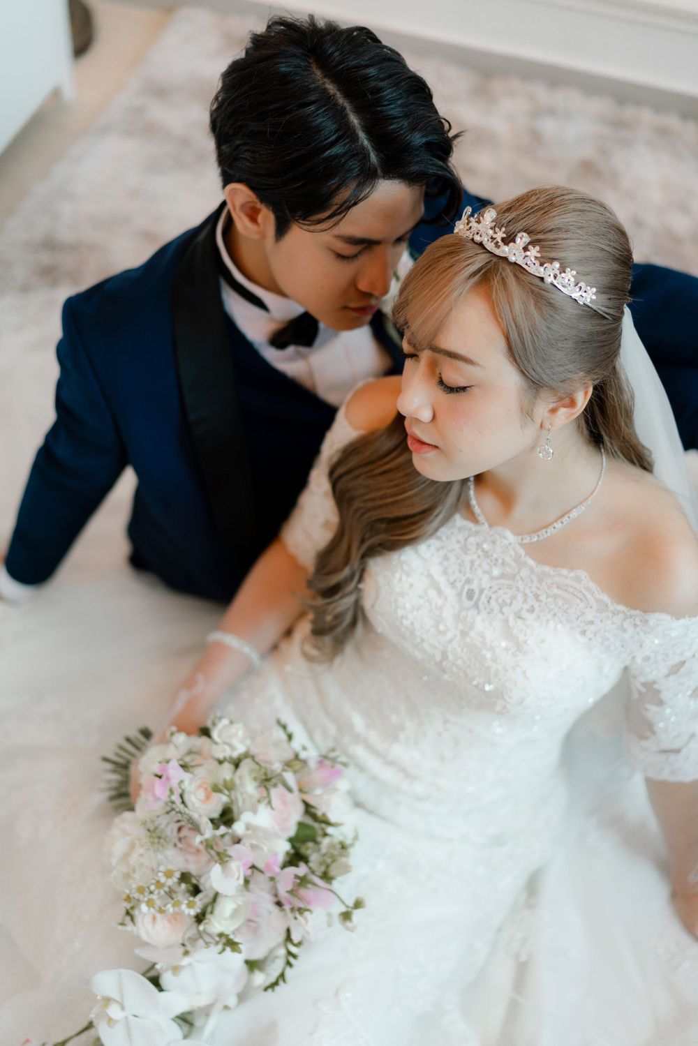 A romantic wedding portrait features a couple in navy suit and white lace gown with delicate pink and white bouquet.