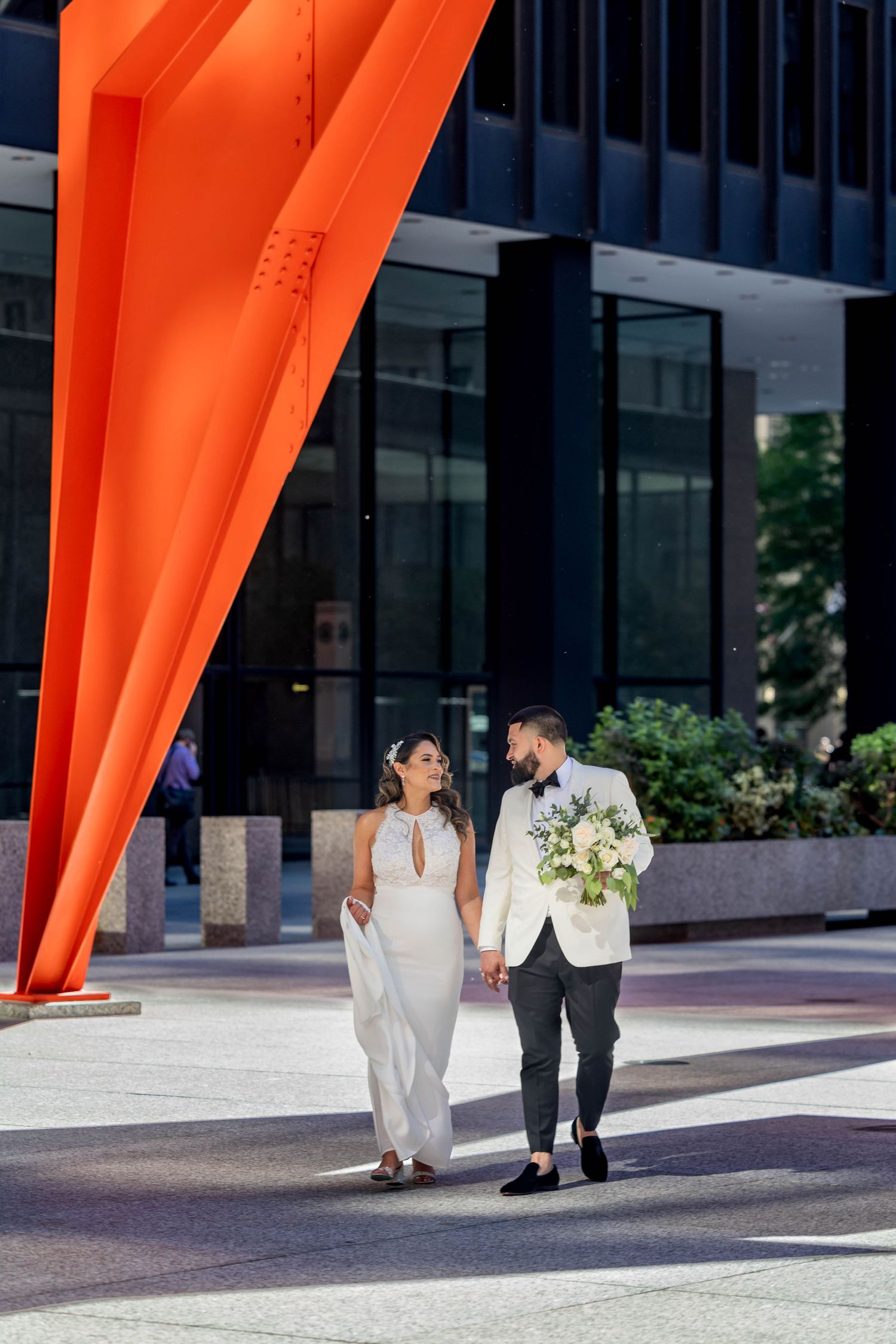 A couple walking under a striking orange sculpture in an urban setting.