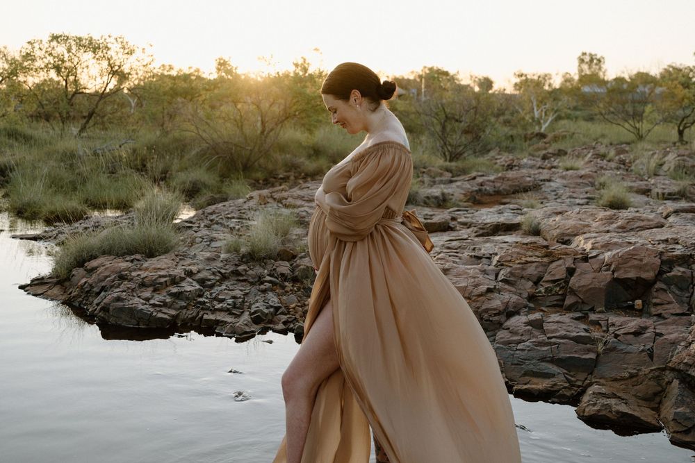 A figure in a flowing rose-colored dress walks barefoot along rocky coastal terrain during a misty sunset.