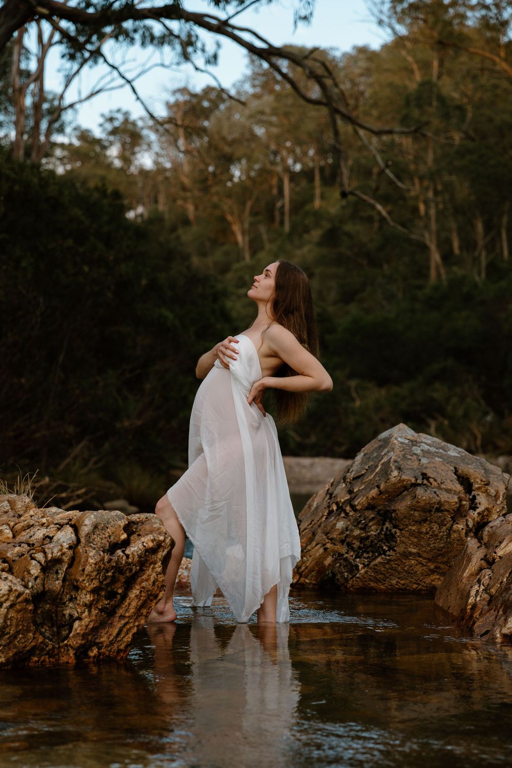 A figure in a flowing white dress poses gracefully on rocks in a stream with a forest backdrop at dusk.
