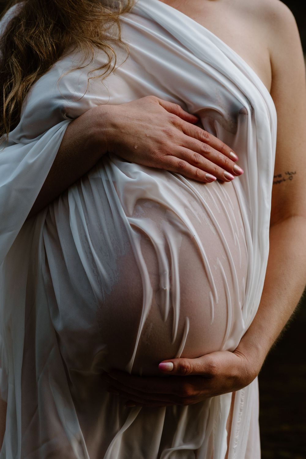 Hands gently caressing a pregnant belly draped in flowing white fabric, creating an ethereal maternity portrait.