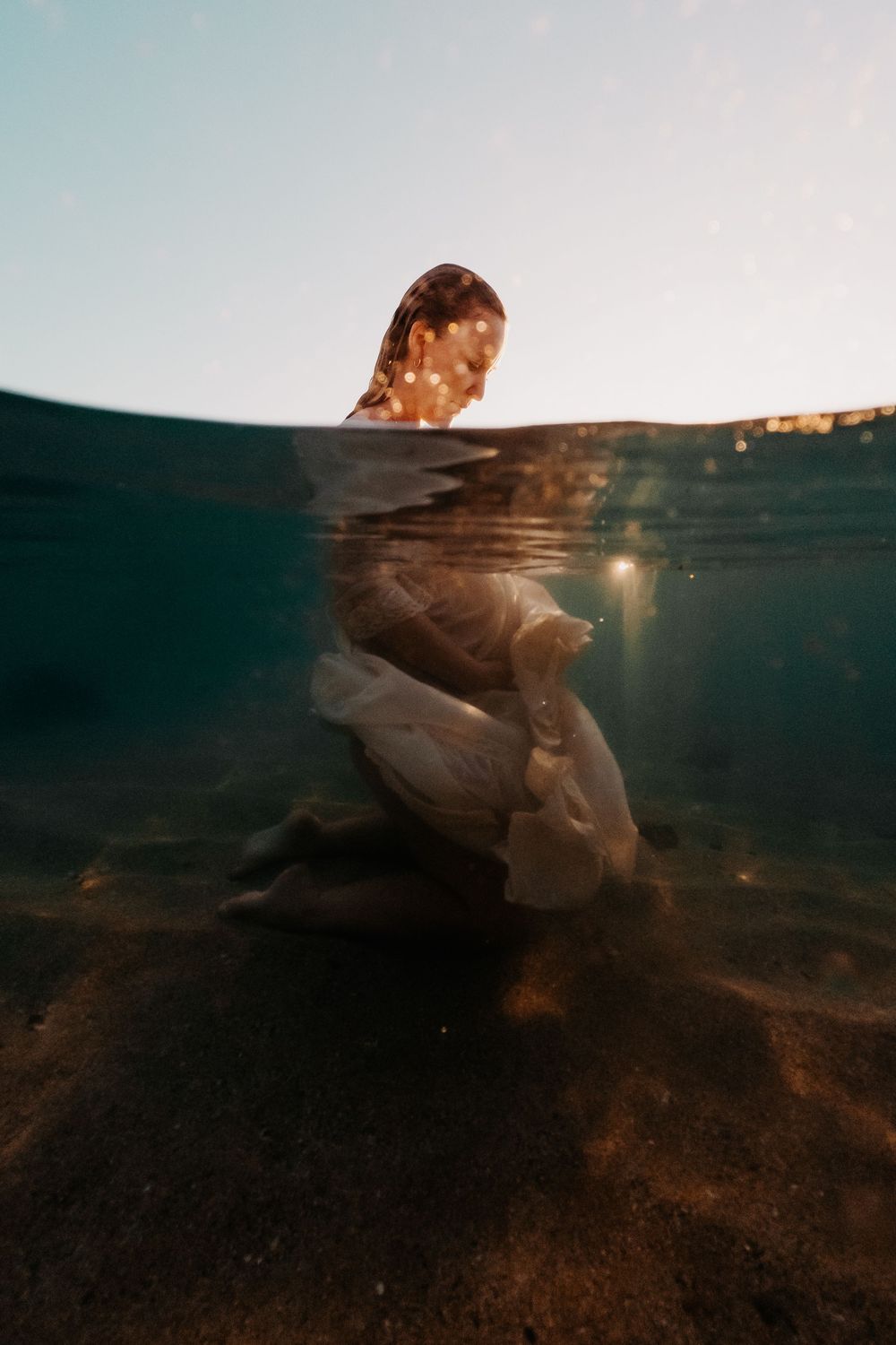 Split underwater photo of a person in white clothing submerged beneath the surface with sunlight filtering through.