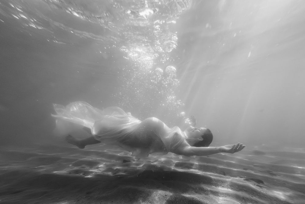 A person floats gracefully underwater as sunbeams pierce through the surface creating an ethereal black and white scene.