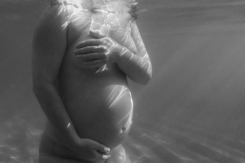 Black and white underwater maternity photo showing a pregnant woman's silhouetted torso cradling her belly.