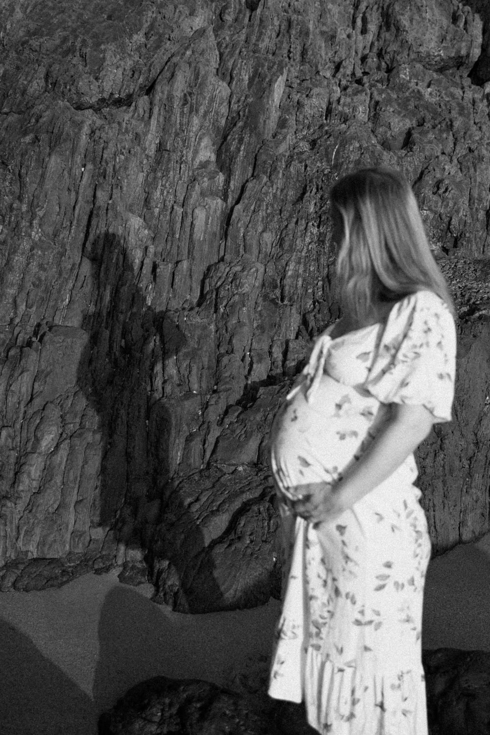 A black and white photo of someone in a floral dress standing against a rocky cliff face in shadow.