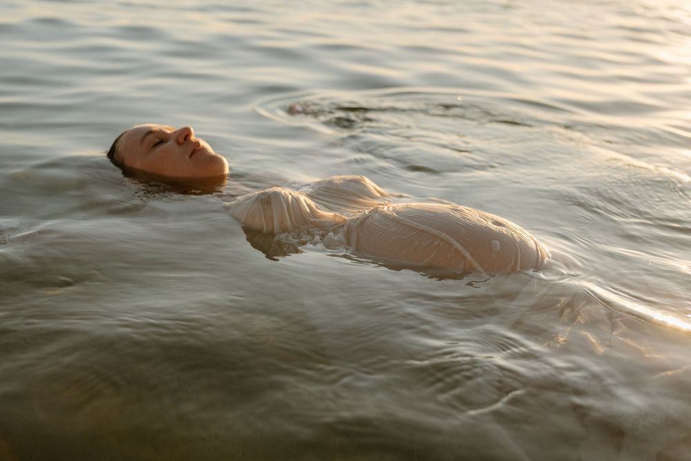 Figure in white dress floats serenely in calm water during sunset with closed eyes.