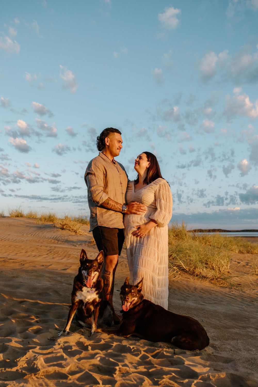 A couple stands on a beach at sunset with their two dogs while fluffy clouds fill the blue sky above.