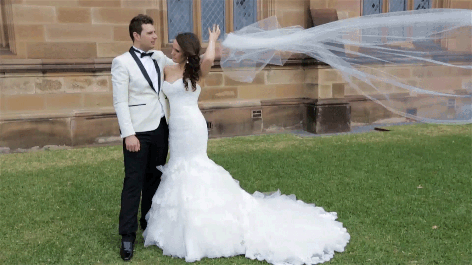 A young married couple standing on green grass next to a sandstone wall. The bride's veil flies away in the wind.