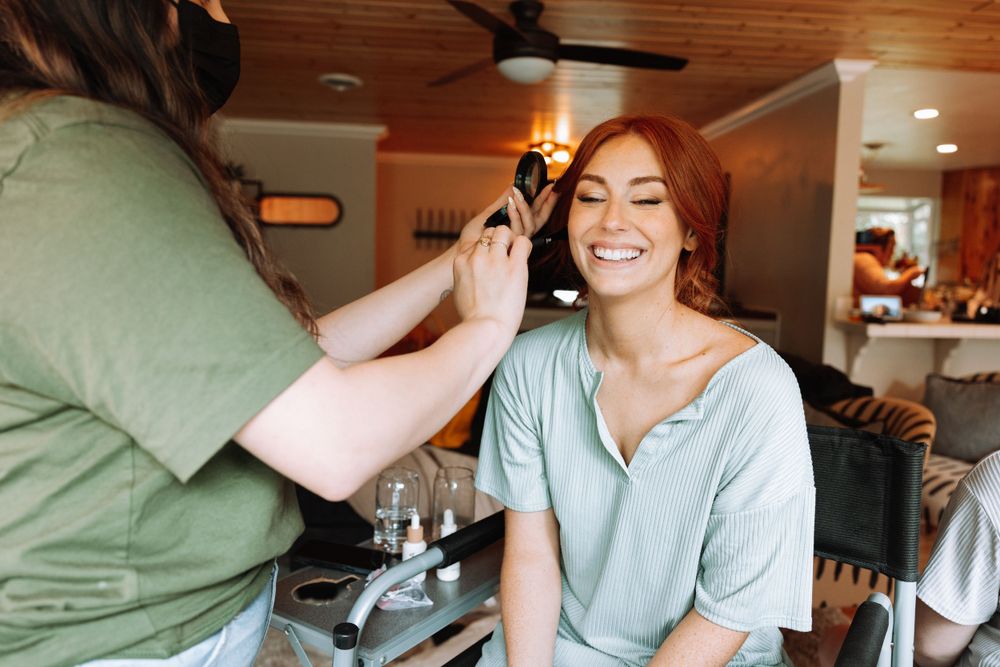 Someone having makeup applied while sitting in a chair and smiling during wedding preparation.