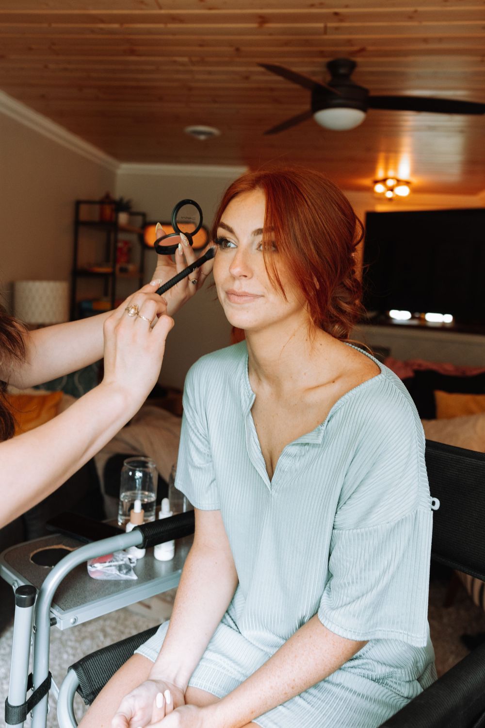 A makeup artist applies cosmetics to a woman wearing a light blue top in a warmly lit indoor setting.