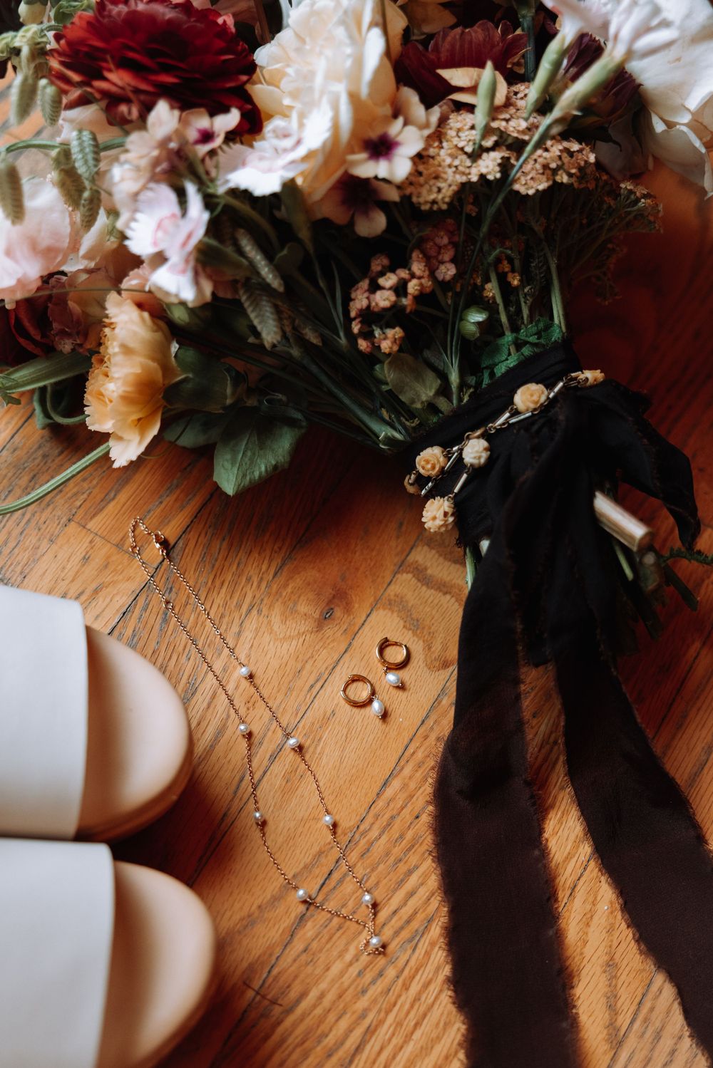 Wedding rings and flowers with black ribbon lie on a wooden surface beside white shoes.