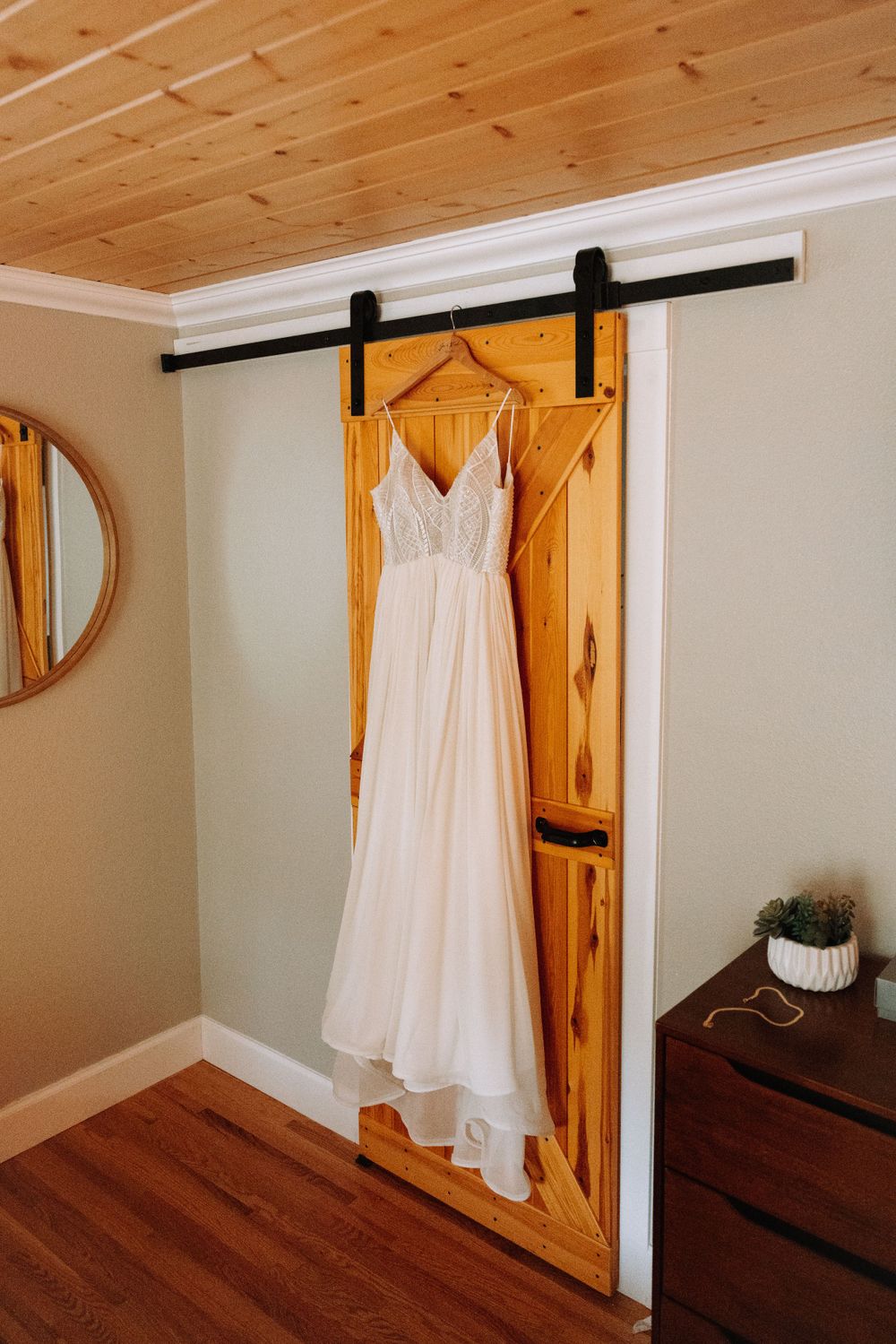 A white lace wedding dress hangs on a rustic wooden barn door with black hardware against a light gray wall.
