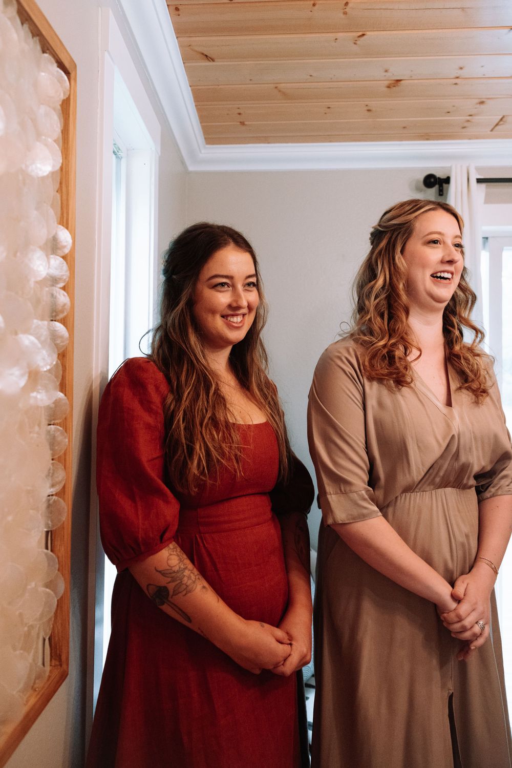 Two women stand smiling in a rustic room with wooden ceiling and string lights, wearing elegant dresses in red and beige.