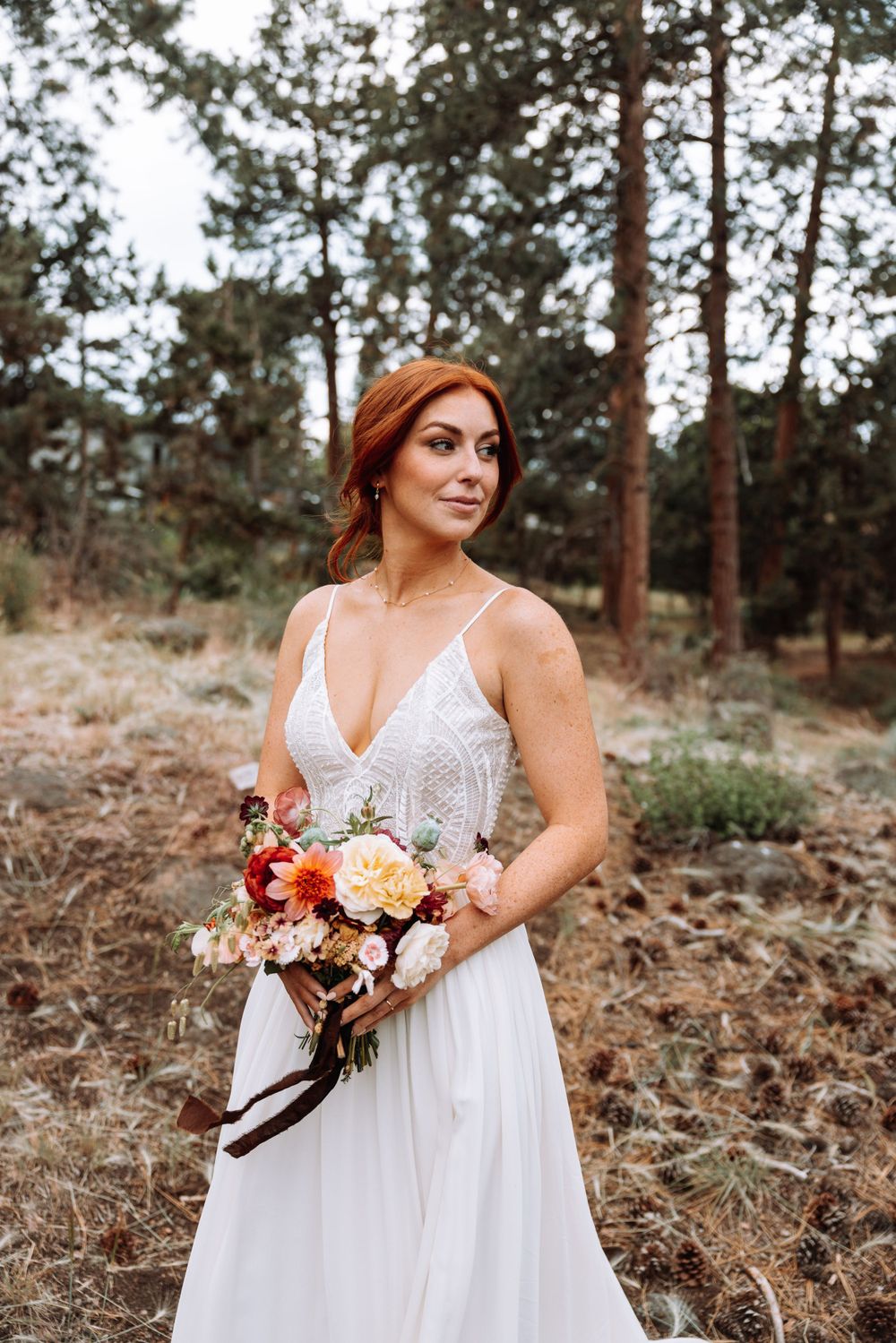 A bride in a white lace wedding dress holds an autumn-colored bouquet while standing in a pine forest setting.