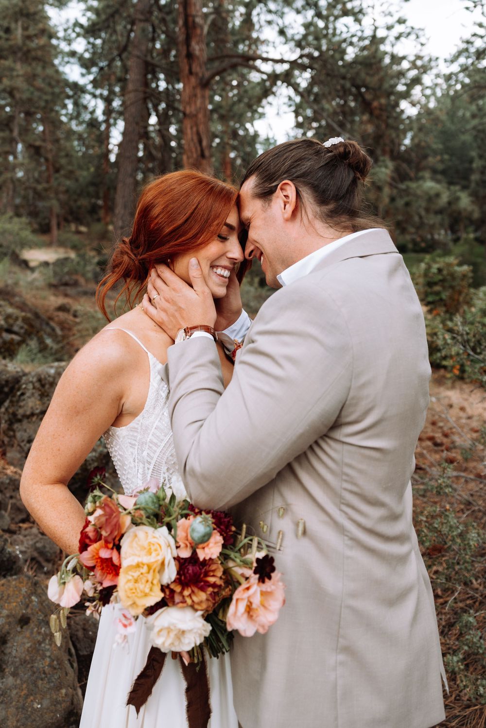 A romantic wedding couple embraces in a forest setting with a colorful autumn bouquet of roses and peonies.
