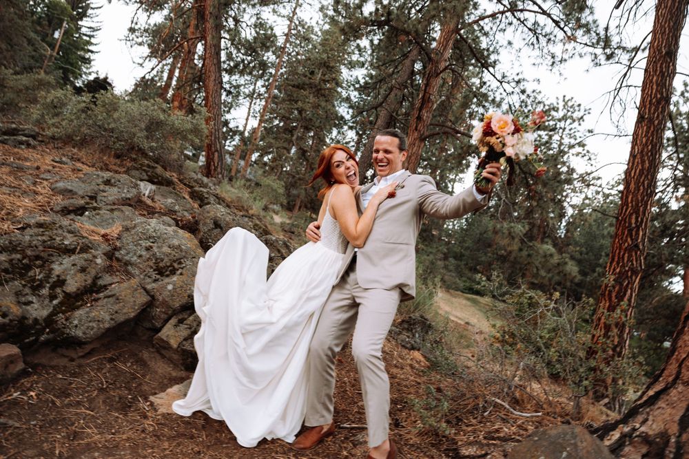 A couple celebrates joyfully among towering pine trees on a rocky forest trail during their outdoor wedding ceremony.