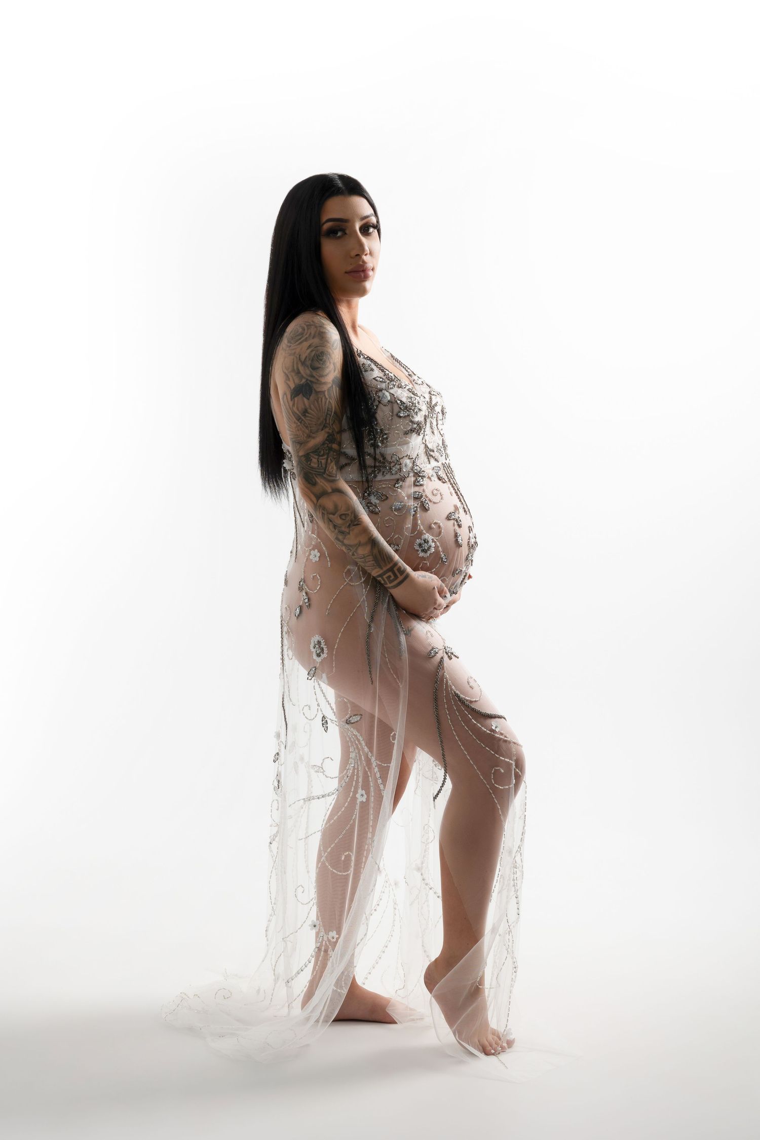 A maternity portrait showing a person in a sheer lace dress posing against a white background.