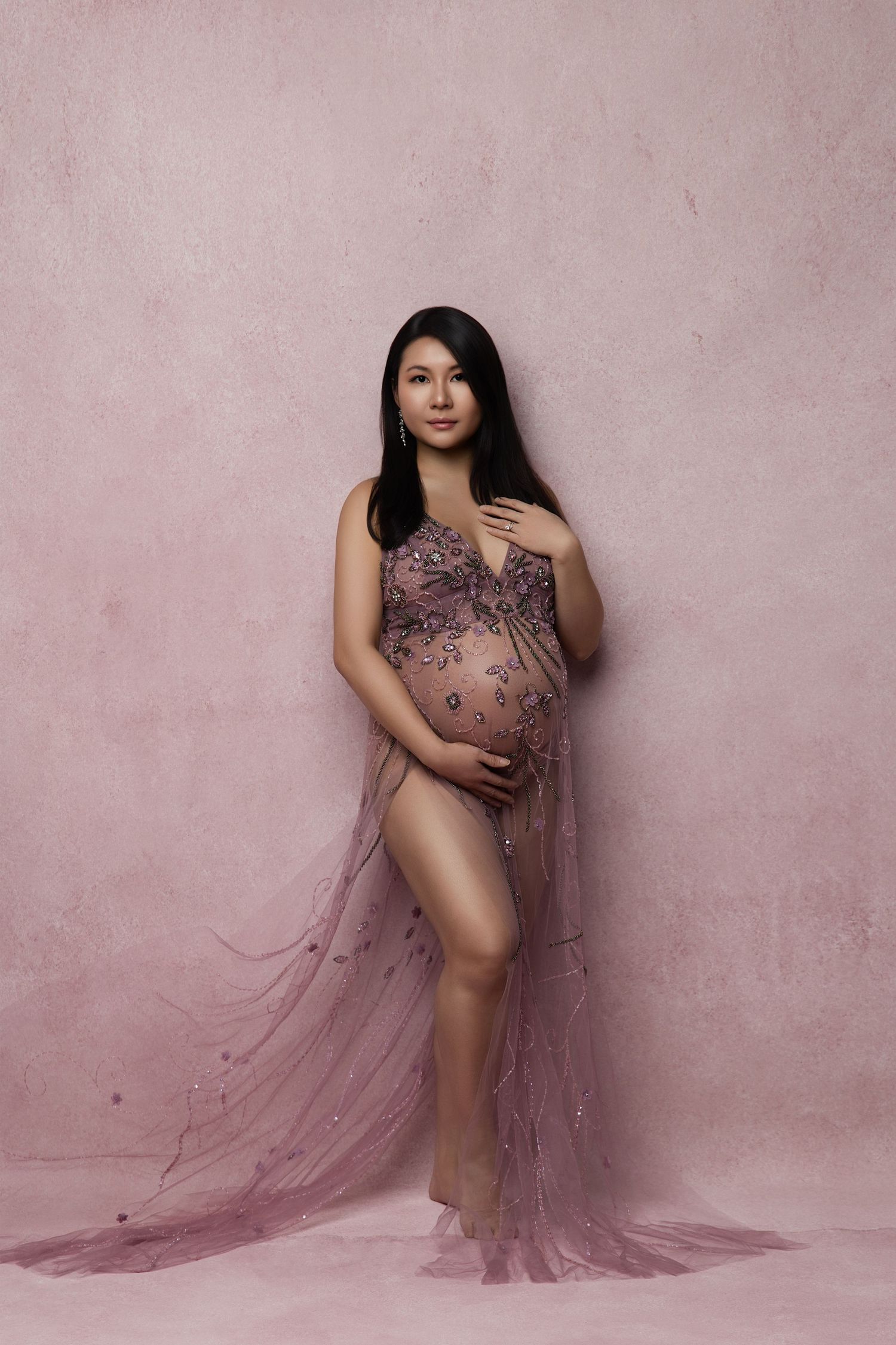 A pregnant woman poses in a sheer mauve maternity gown against a pink textured backdrop for an elegant portrait.