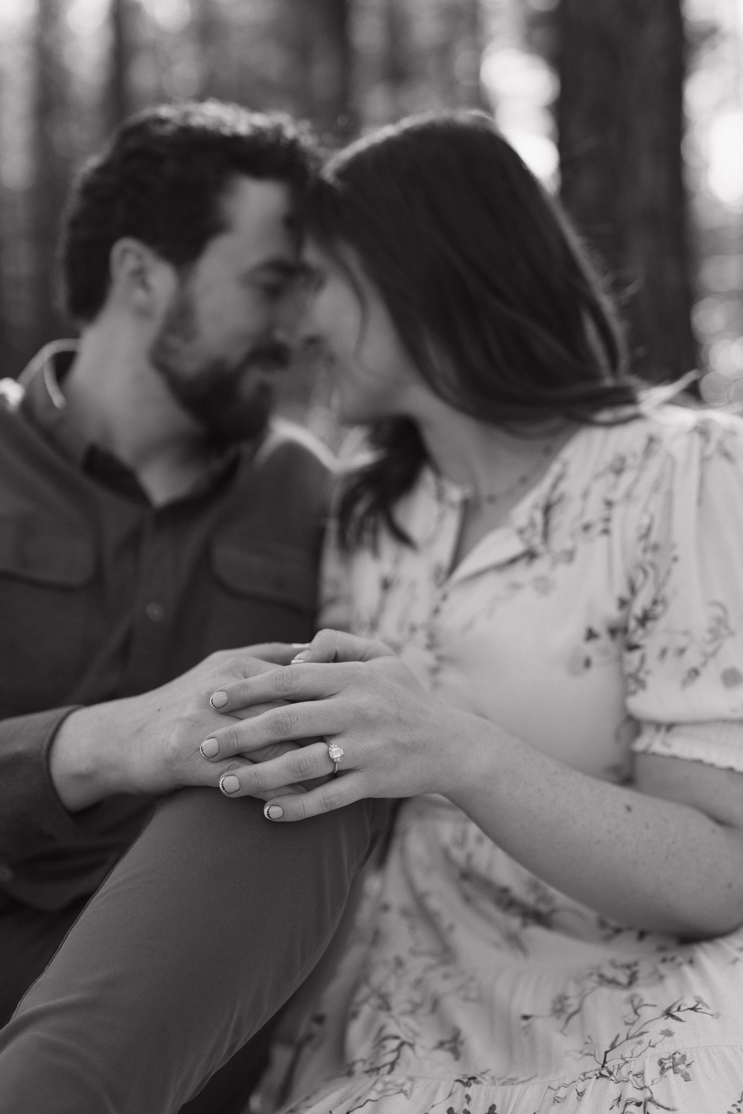 Black and white photo of couple almost kissing, woman displaying engagement ring on hand in forest setting.