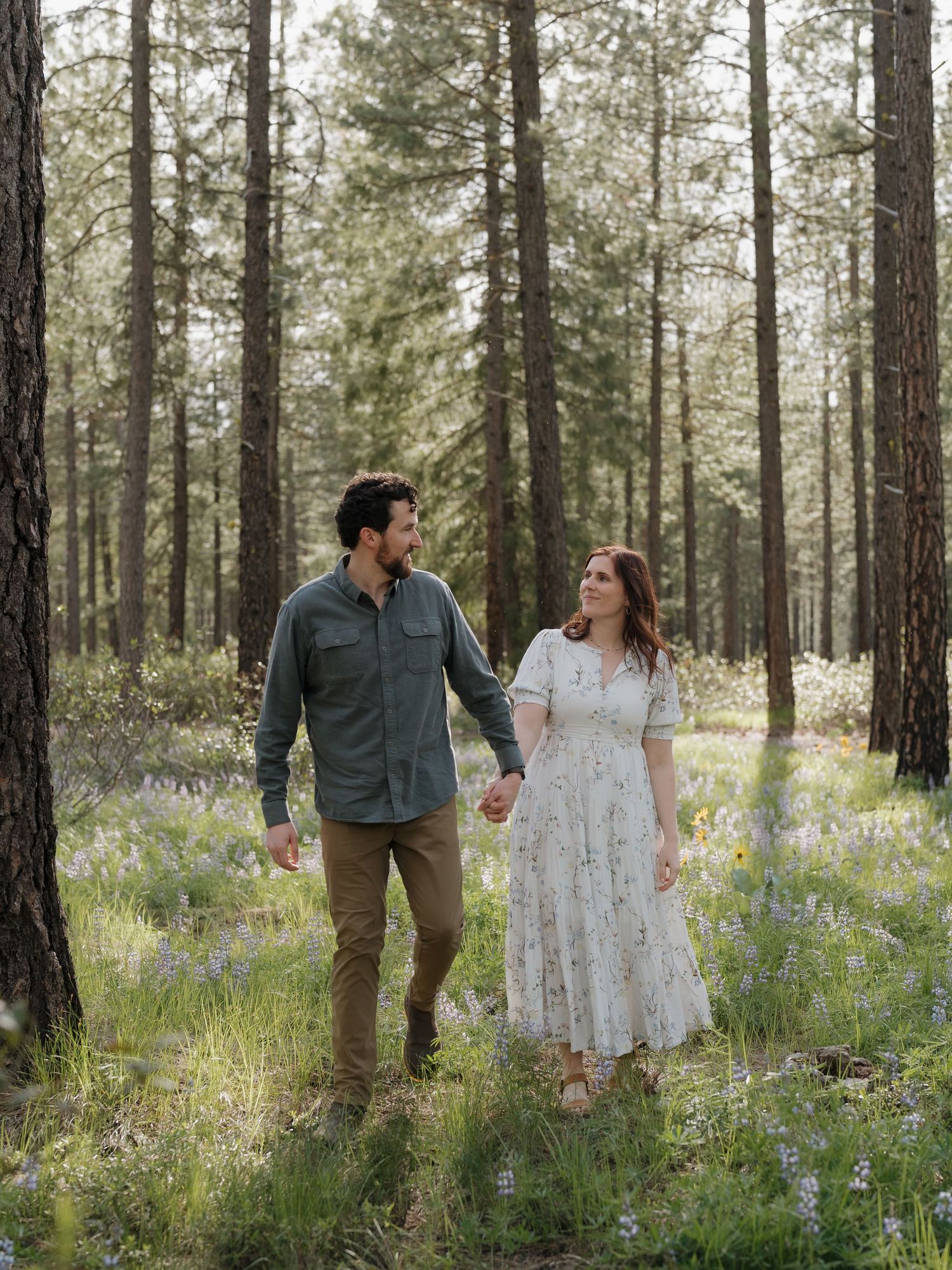 Couple holding hands walking through a sunlit forest meadow with wildflowers, man in green shirt and woman in floral dress.