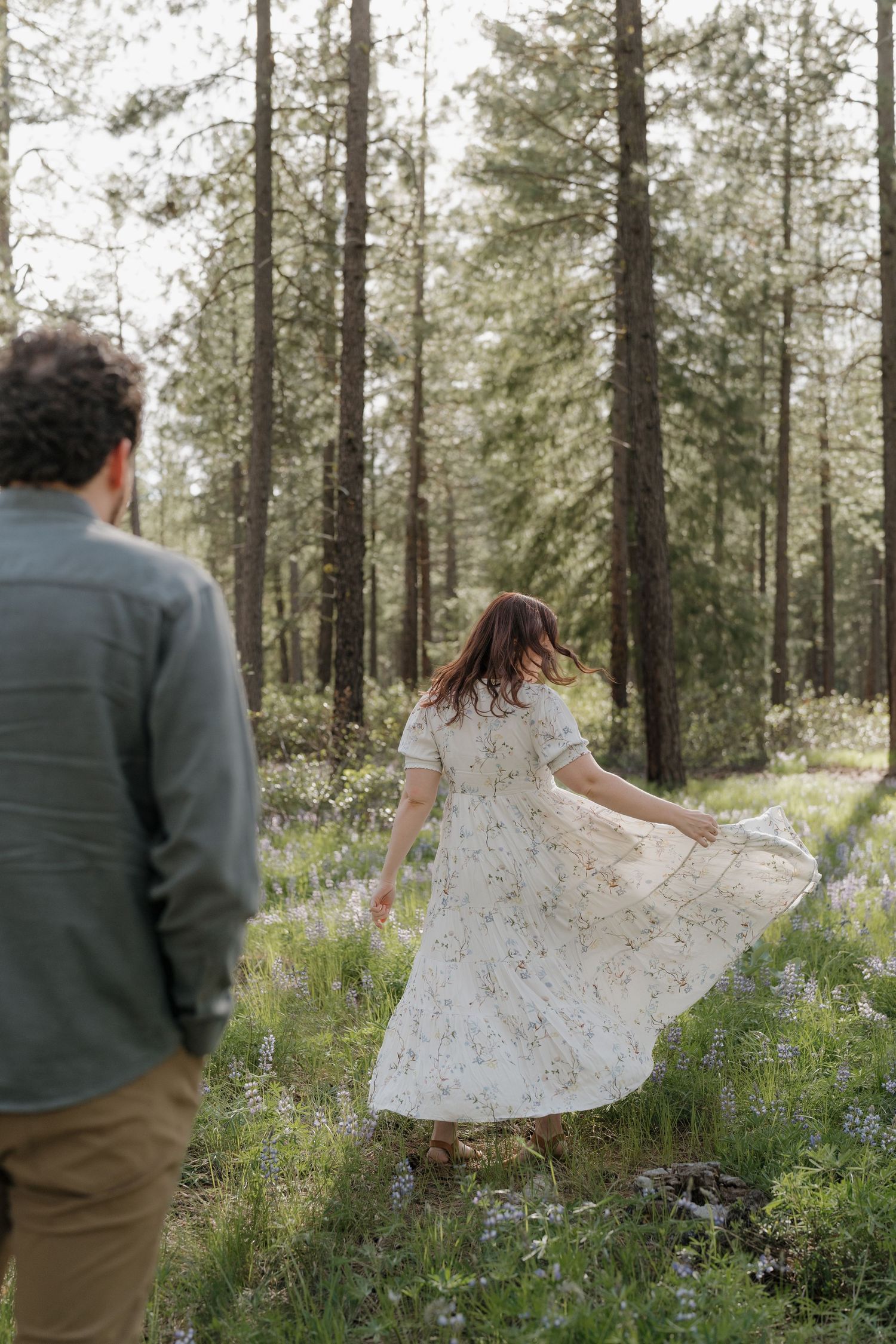 Woman in floral dress twirling in a sunlit forest clearing with tall pine trees and wildflowers.