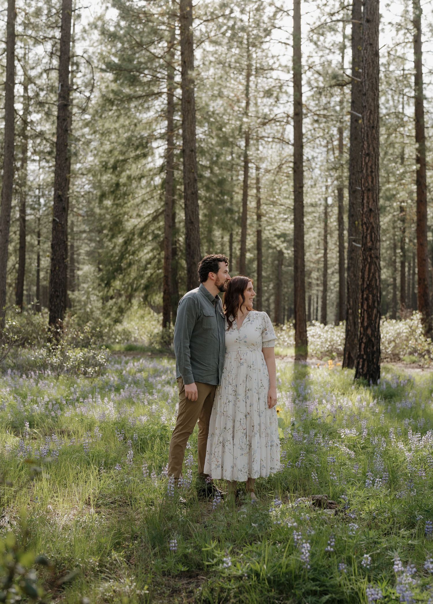 Couple standing among wildflowers in a pine forest, woman in white dress, man in gray jacket, sharing a tender moment.