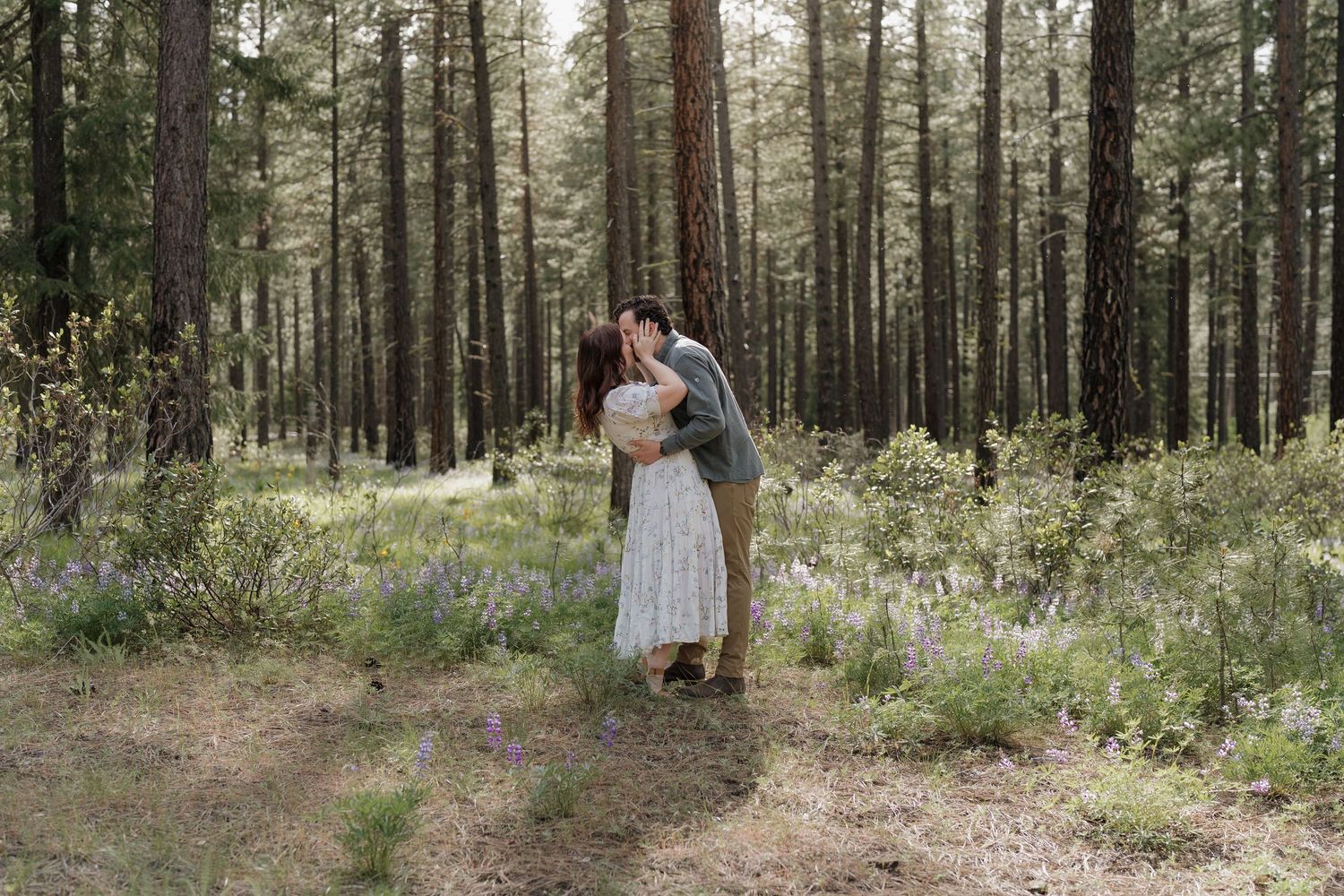 Couple sharing a romantic kiss in a sunlit pine forest with wildflowers blooming around them.