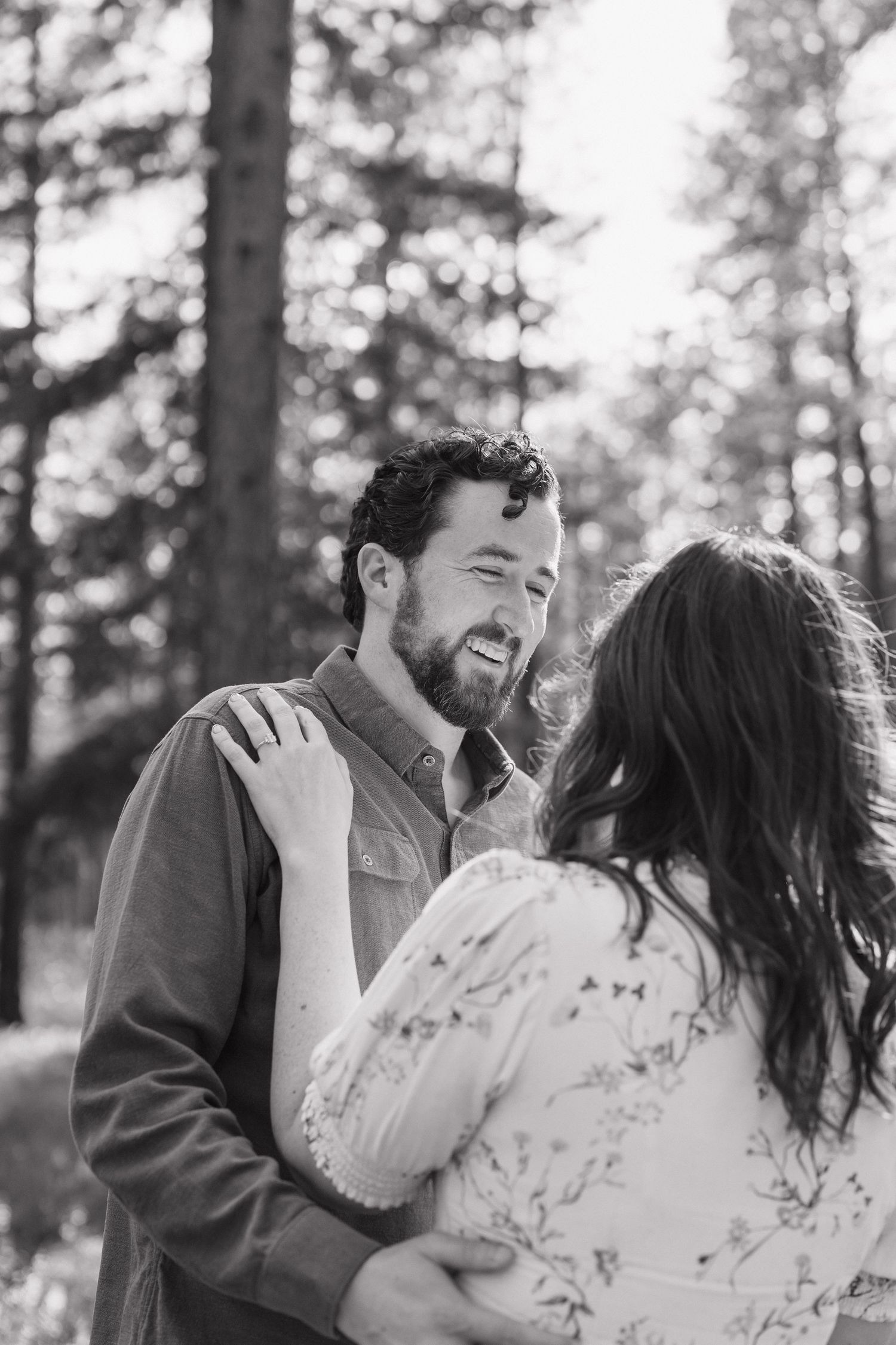 Black and white photo of a bearded man smiling at a woman during a romantic forest engagement session.